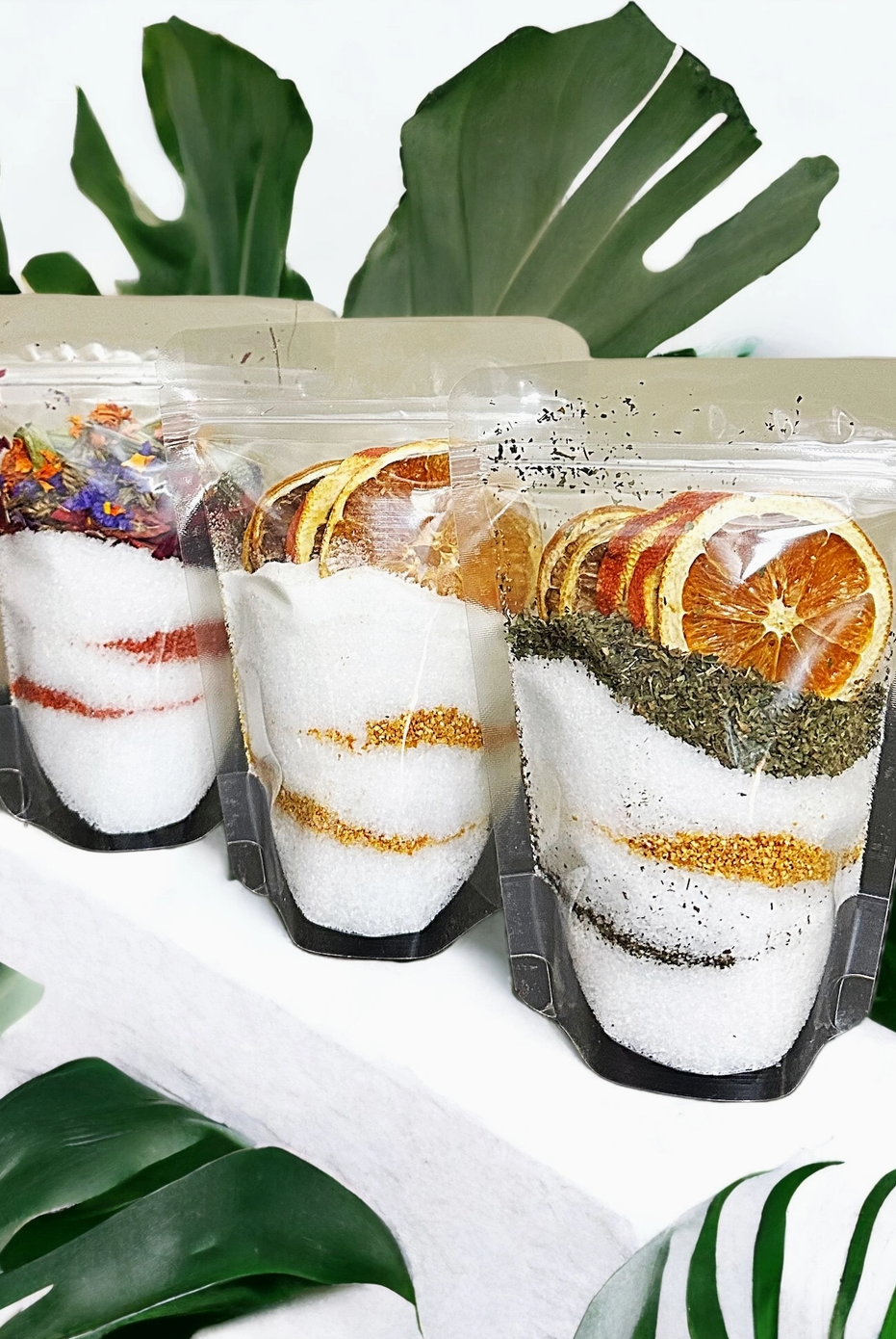 Three packages of bath salt with dried fruit and herbs on a white surface with green leaves in the background