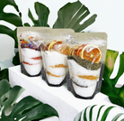 Three packages of bath salt with dried fruit and herbs on a white surface with green leaves in the background